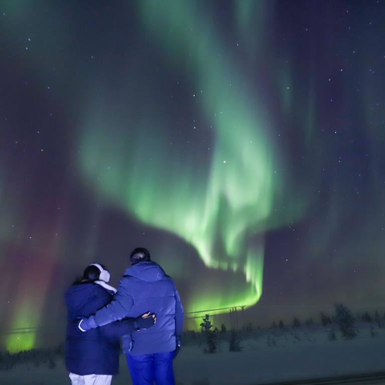 Couple standing arm in arm watching the Northern Lights over a snowy landscape