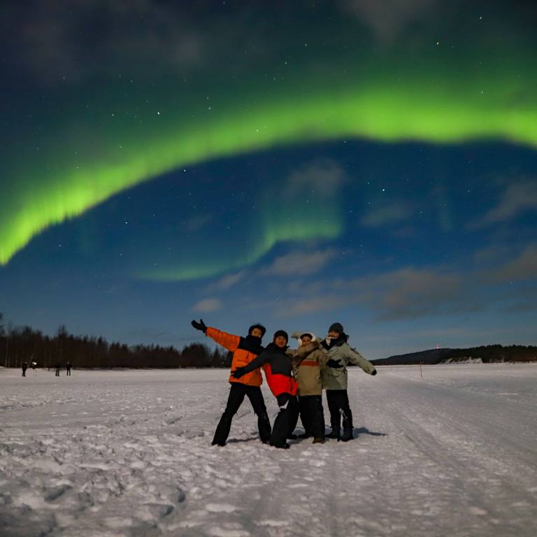 Group of four people celebrating on a frozen lake under a green Northern Lights arc