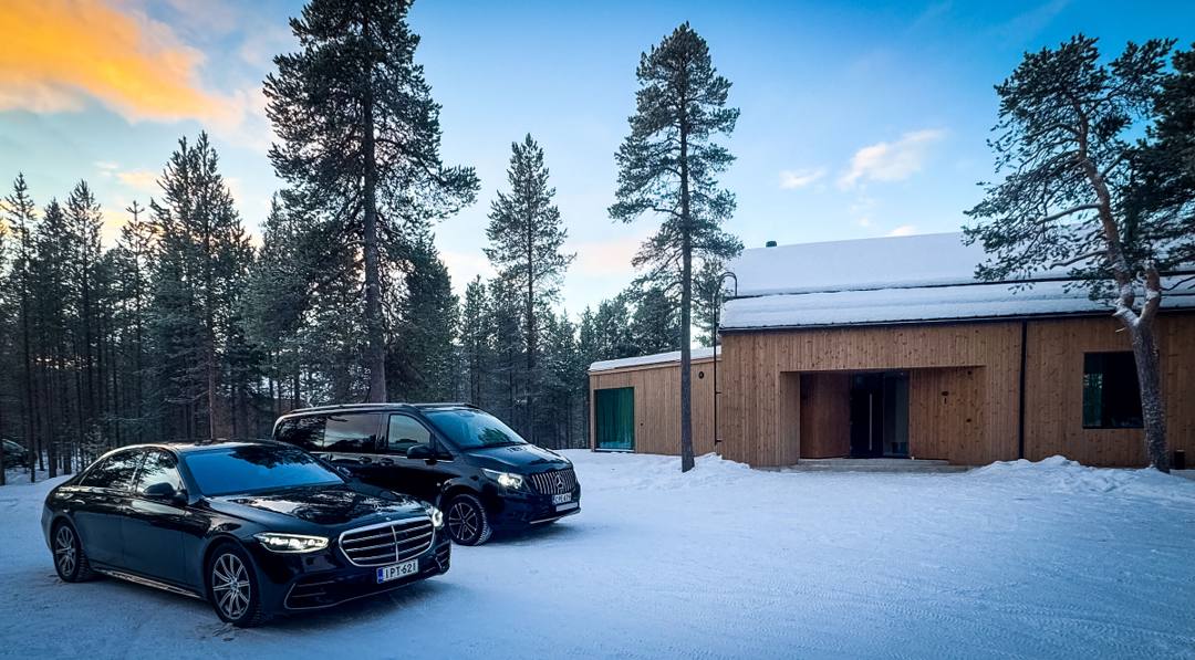 Black Mercedes-Benz sedan and black Mercedes-Benz van outside a wooden cabin among snowy pine trees in Lapland