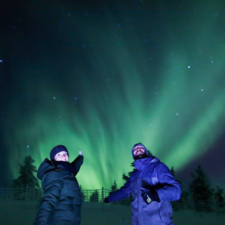 Two guests in winter jackets looking up at bright Northern Lights